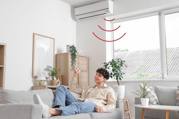 Junger Erwachsener liegt fröhlich auf Sofa und bedient eine Klimaanlage per Fernbedienung